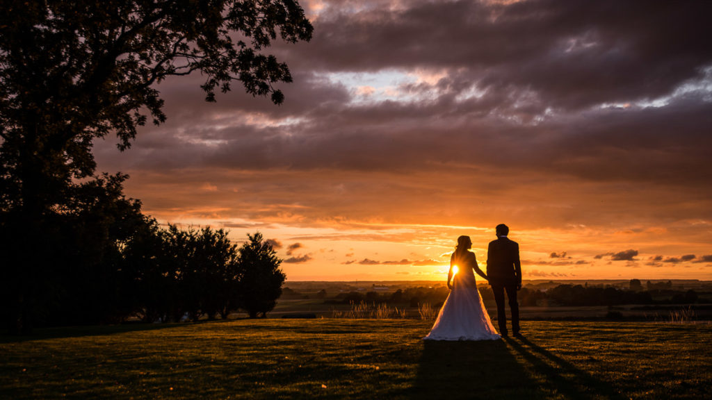 Tones of orange summer sunset with bride and groom stood side by side gazing at each other, the sun just peeking to the side of the bride
