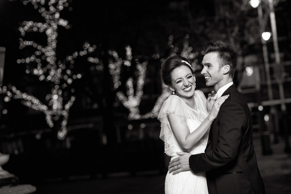 Black and white image with fairy light adorned trees in the background as bride and groom laugh with each other in the foreground