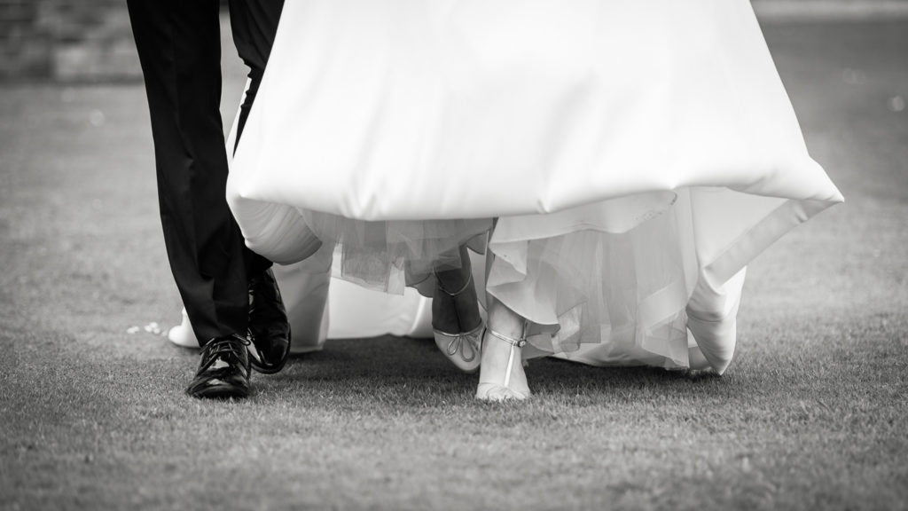 Black and white image focussed closely on the bride and groom's feet as they walk across the grass with bride lifting her dress