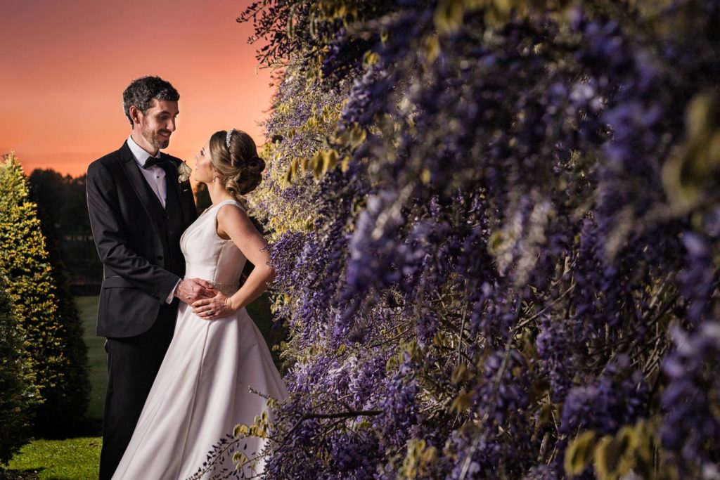 Bride and groom posed next to purple wisteria in full bloom with sunset background