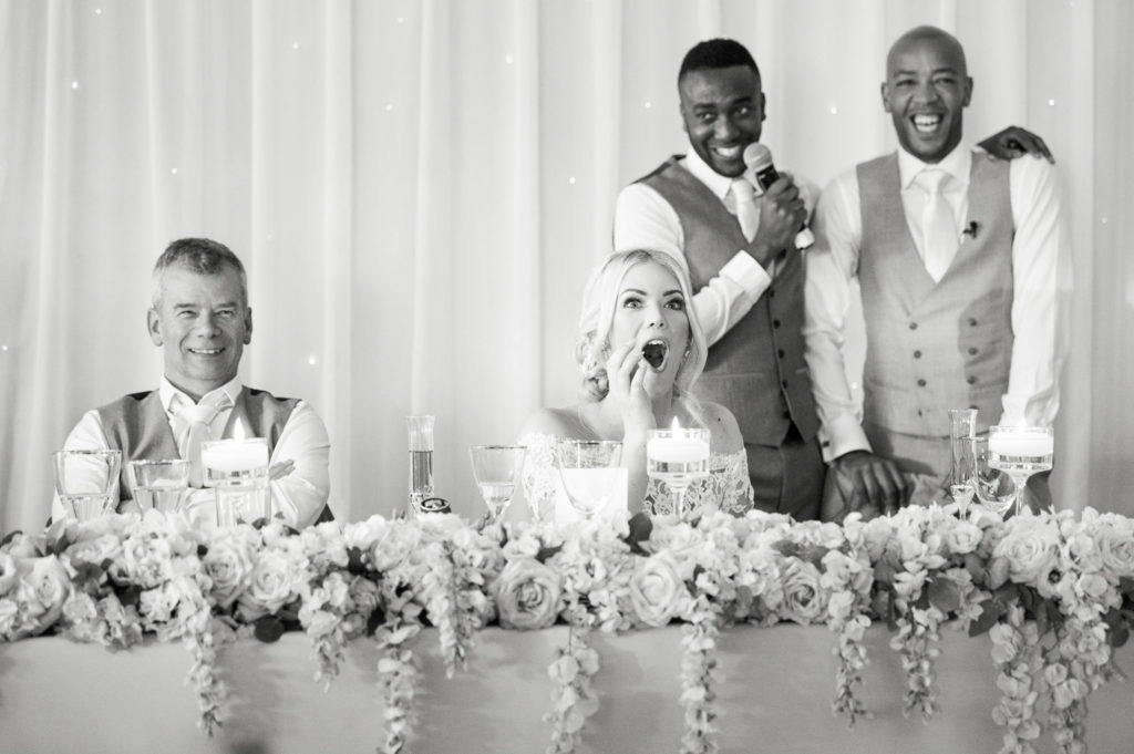 Bride agog during Best man speech with groom stood behind her laughing