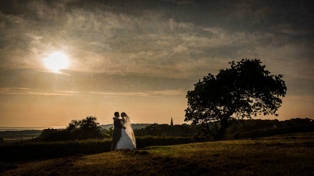 Milky pale sunset over the Wirral Estuary while bride and groom hold in an embrace in a field