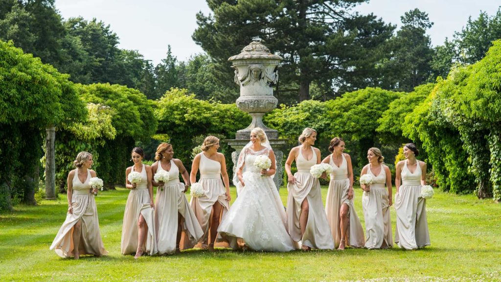 Bride and eight bridesmaids walking across the rear lawns at Thornton Manor in white and nude tones with backdrop of luscious greenery