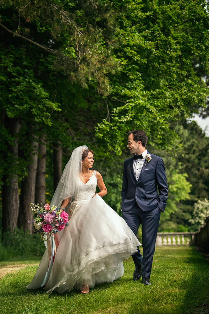Bride walking holding dress in one hand and vibrant bouquet in other with groom by her side hands in pockets of tux