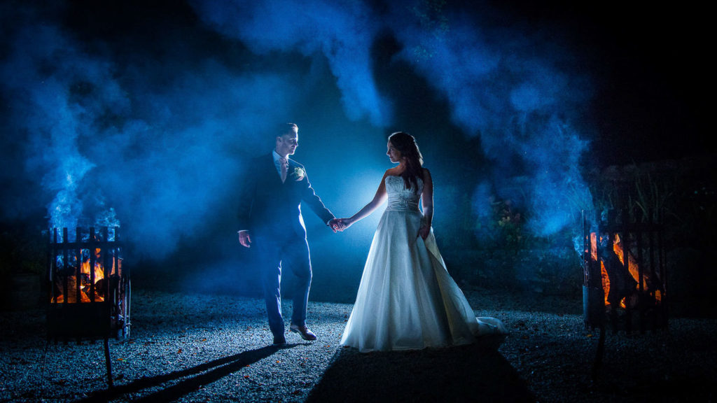 Bride and groom hand in hand with fire pits burning orange on either side and smokey deep blue backdrop