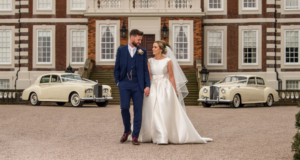 Newlyweds walking along the drive at Knowsley Hall with cream Rolls Royce cars parked behind them