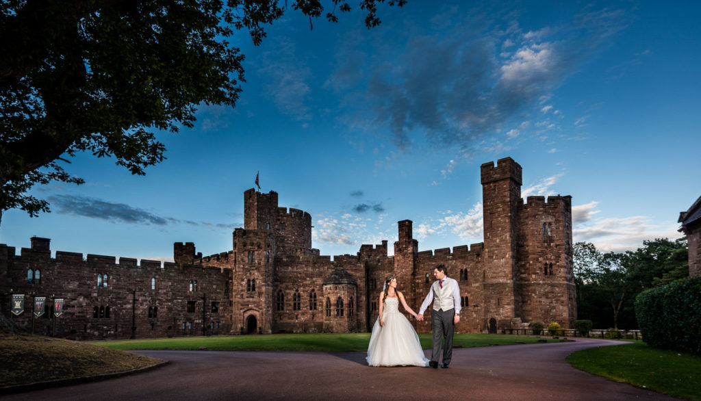 Newlyweds walking along path hand in hand at Peckforton Castle with summer evening blue sky overhead