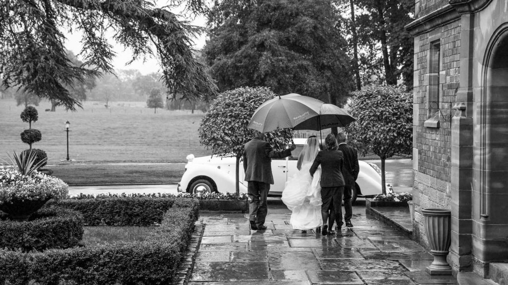 Bride walking away from camera towards wedding car in pouring rain with Colshaw Hall staff holding umbrellas over and scooping up her dress