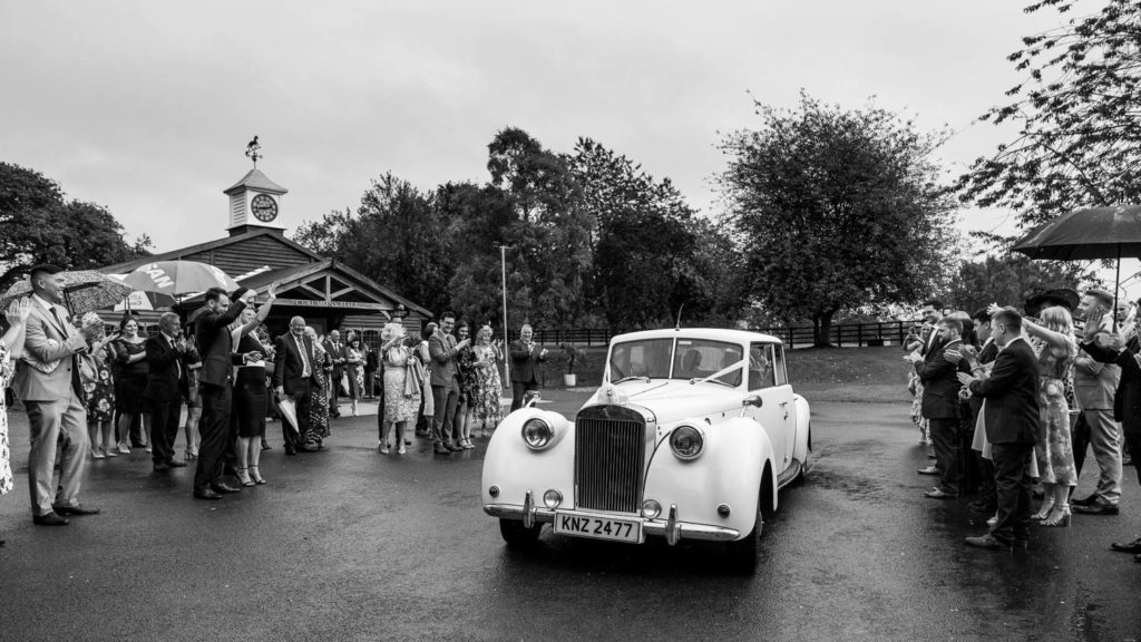 Bride and groom in wedding car at Colshaw Hall after ceremony in the rain with guests waving them off