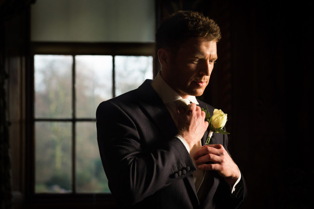 Groom stood in bright window light straightening his tie before the ceremony with cream rose buttonhole