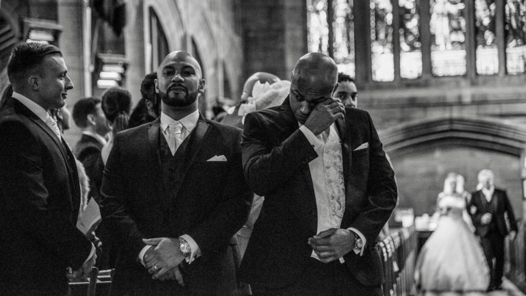 Groom wiping away a tear in foreground as Bride and her father approach down the aisle