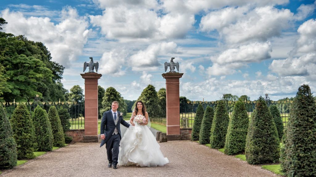 Bride and Groom happily walking up driveway with groom helping to carry dress at Knowsley Hall