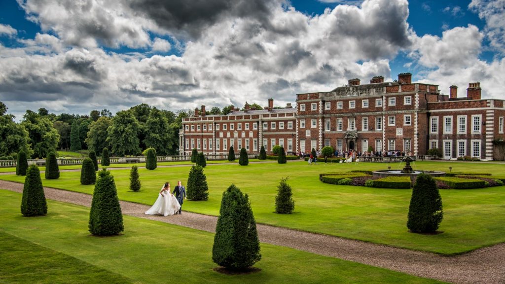 Impressive panoramic featuring newlyweds walking through rear gardens at Knowsley Hall as guests enjoy drinks on terrace