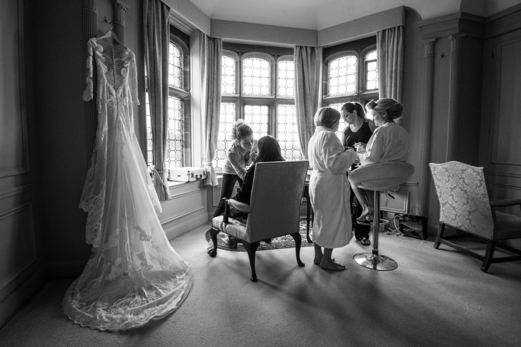 Finishing touches to bridal hair and makeup in bay window with dress hanging up to the side