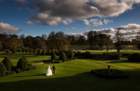 Dramatic panoramic image of bride and groom walking through lush green gardens under a blue sky at Knowsley Hall