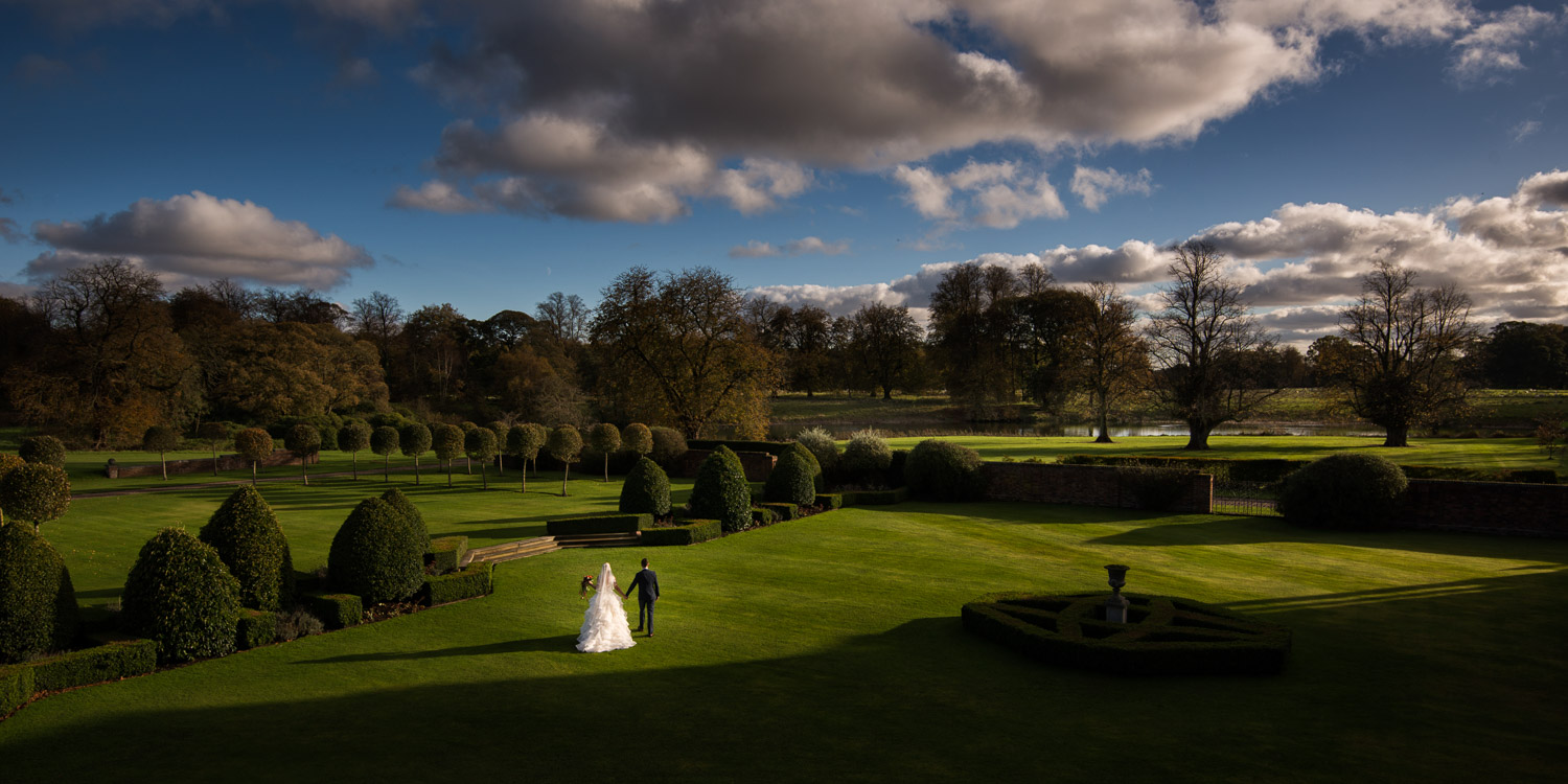 Dramatic panoramic image of bride and groom walking through lush green gardens under a blue sky at Knowsley Hall