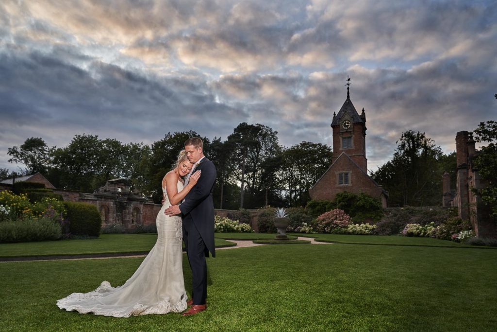 Bride and groom in a gentle hold in the gardens featuring a clocktower behind them