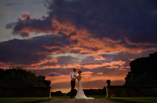 Stunning sunset of red and orange cloud filled sky with bride and groom looking at each other in the middleground