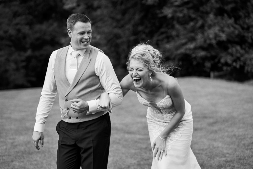 Black and white image of bride and groom walking through the gardens with bride bent over laughing