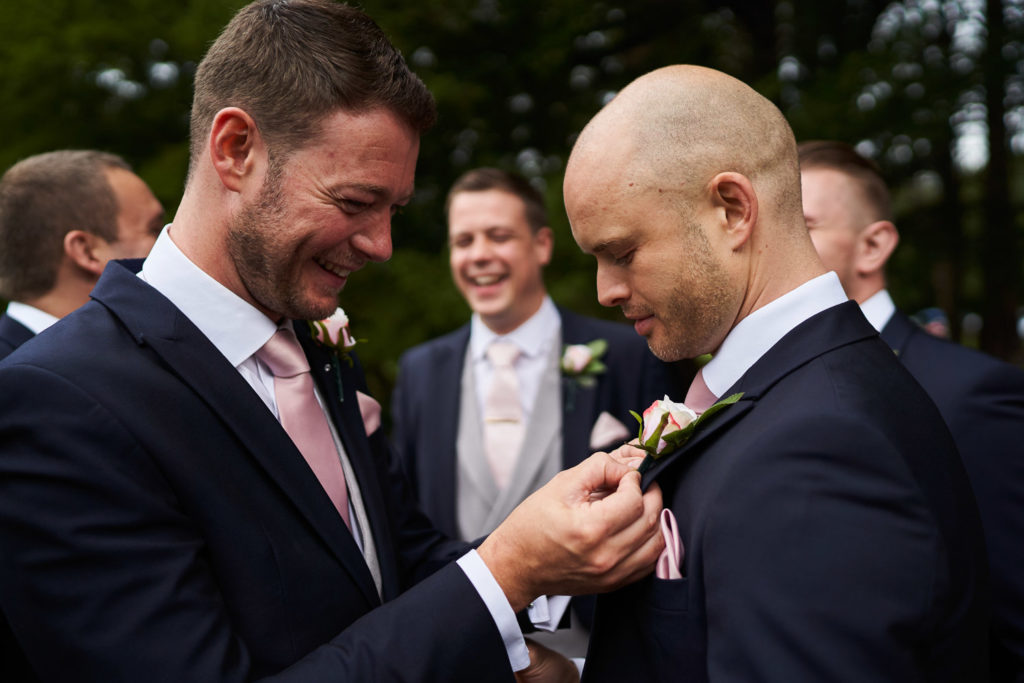 Usher laughing as he pins a pink buttonhole rose to another usher's lapel
