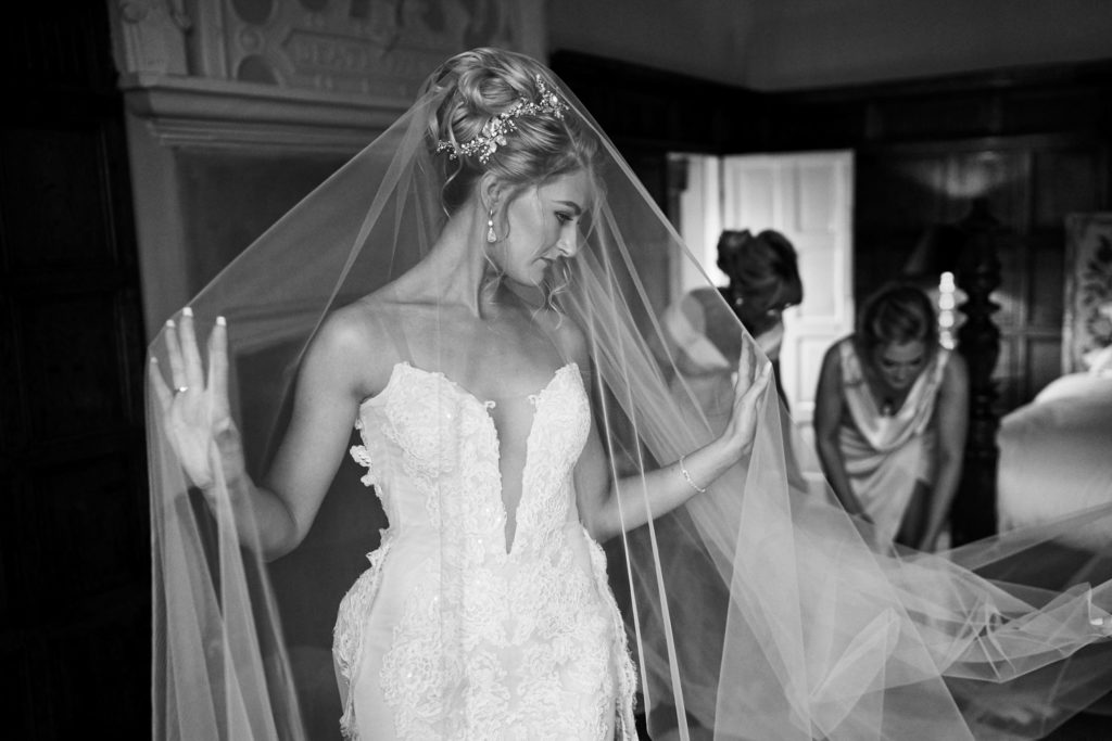 Bride posing under her veil as mum and bridesmaid check length to cover the dress