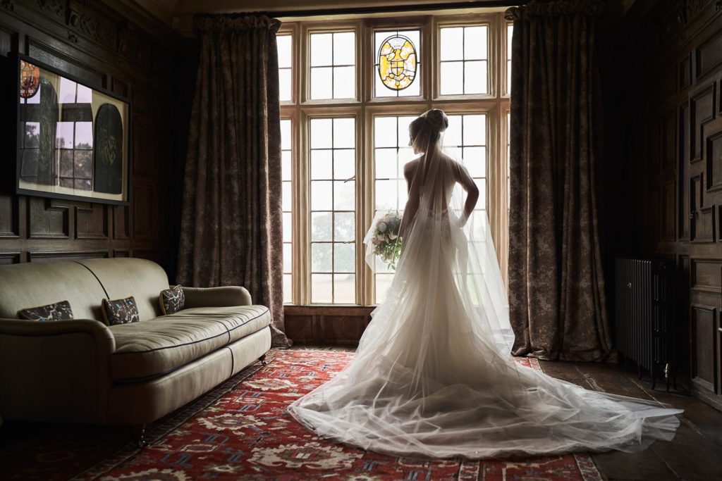 Back shot of bride stood in soft window light with bouquet to her side