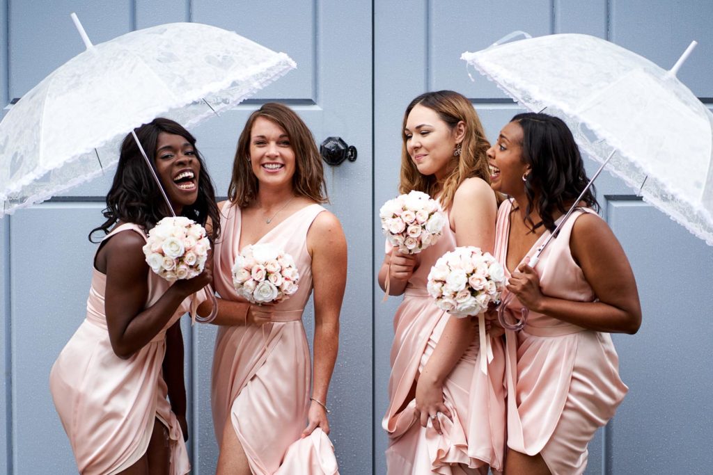Bridesmaids in pale pink stood under pretty umbrellas with dresses hitched up