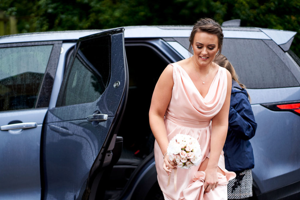 Bridesmaid in pale pink dress with cowl neckline leaving the wedding car