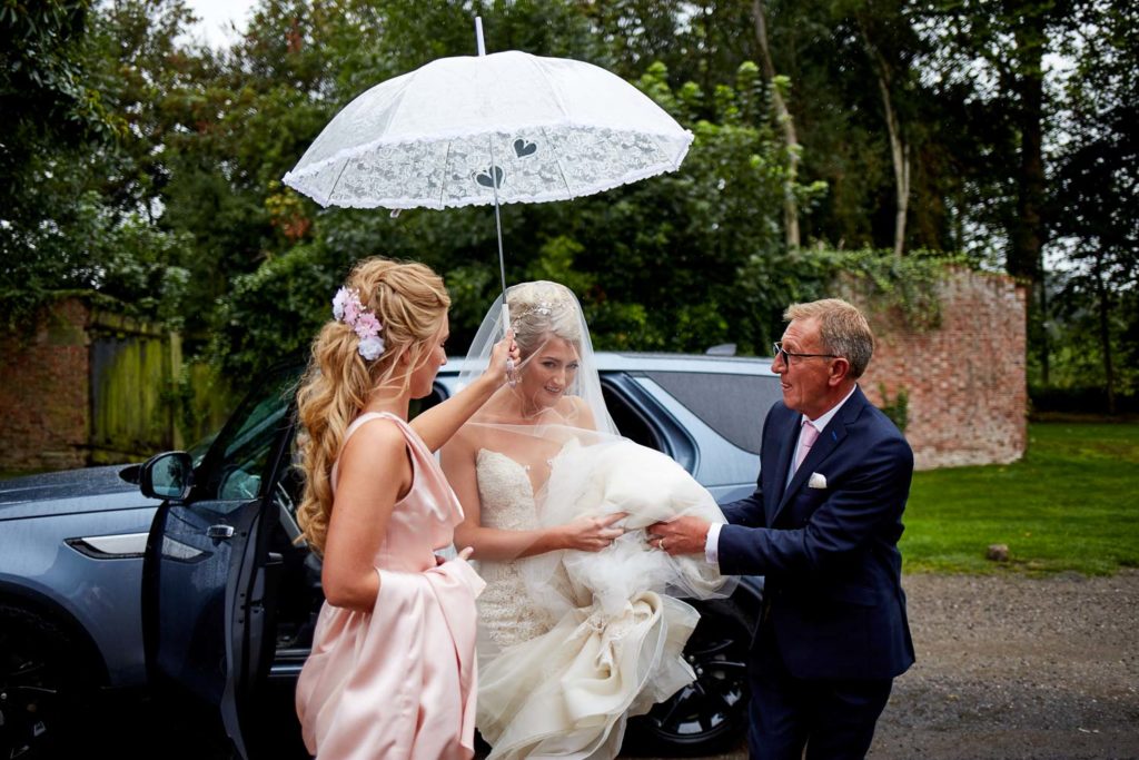 Bride alighting from wedding car as bridesmaid holds an umbrella over her