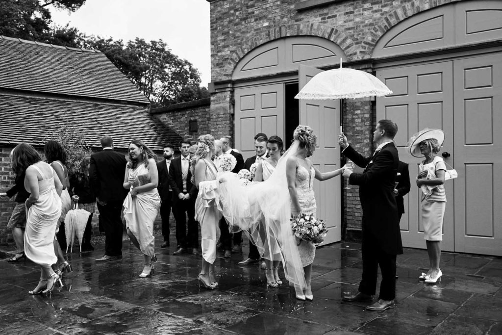 Happy couple leaving the ceremony under an umbrella with bridesmaids lifting bride's dress