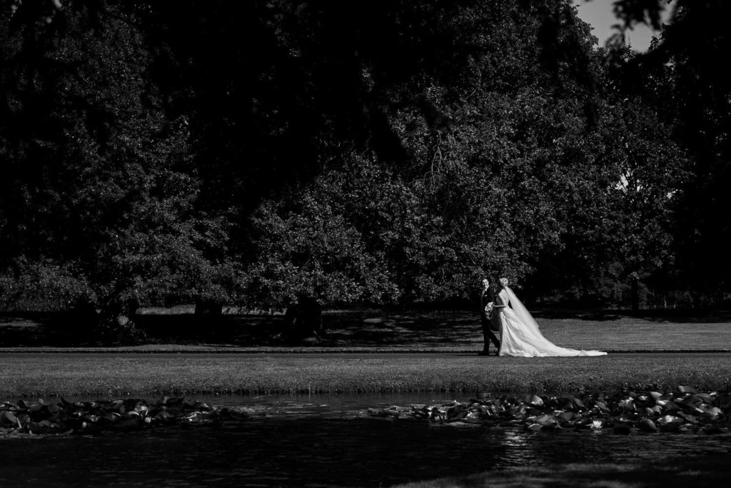 Bride and groom walking on the lawns across the lake at Dorfold Hall in black and white