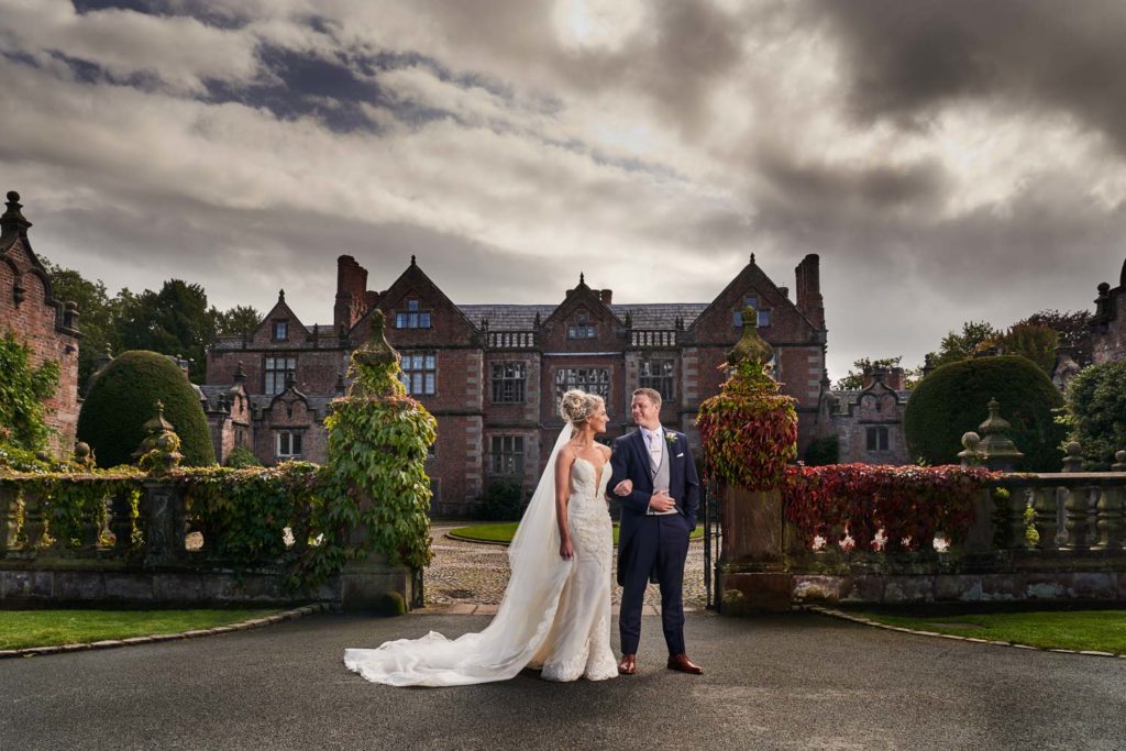 Husband and wife posed in front of Dorfold Hall with muted tones of greens, reds and greys