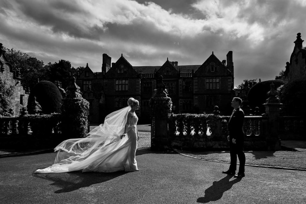 Bride walking towards her husband as her dress and veil billow in the breeze behind her in black and white