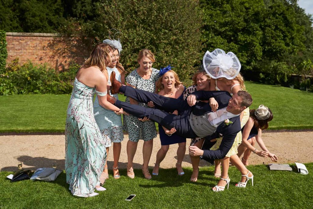 Six female guests laughing while trying to hold the groom lengthways but he is about to hit the floor