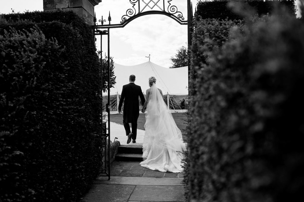 Back shot of newlyweds as they walk through an iron arch in the hedgerow to their wedding breakfast