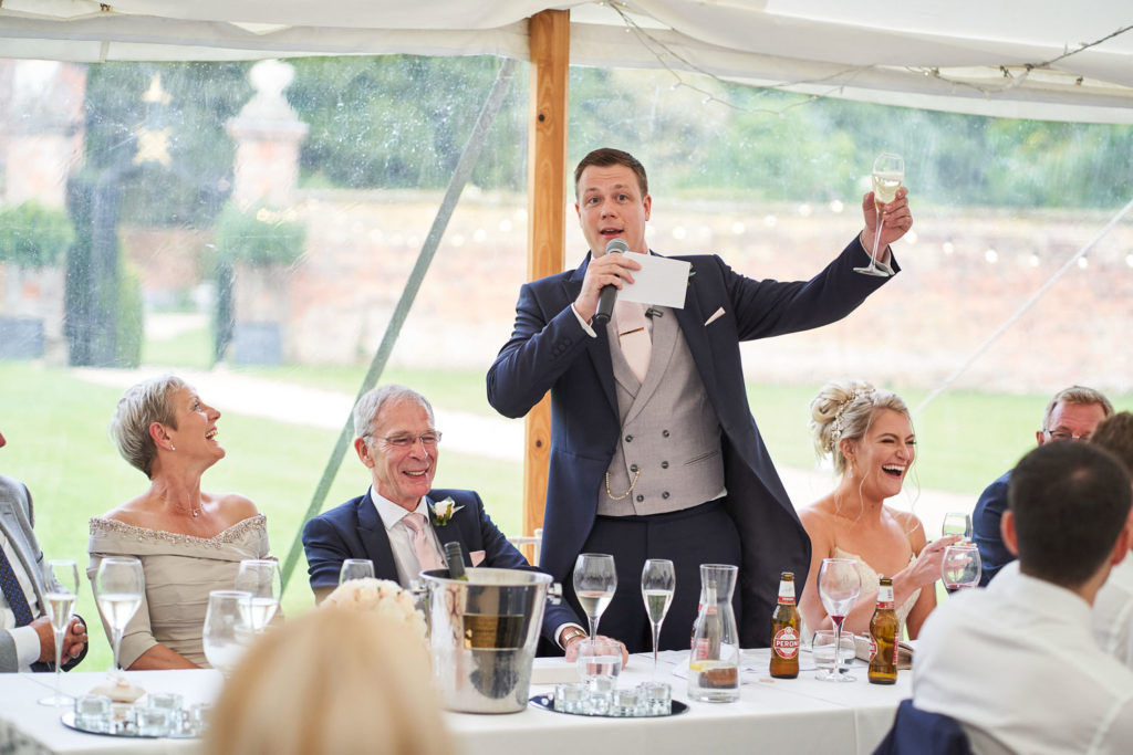Groom raises his glass to propose a toast as top table are all laughing heartily