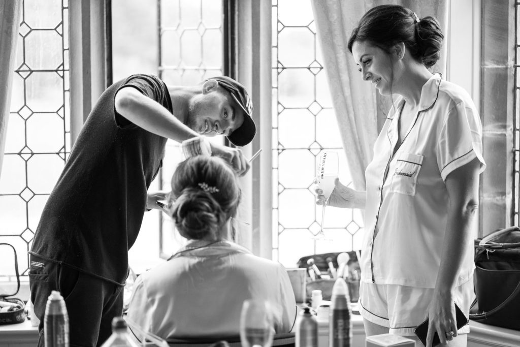 Bridesmaid stood drinking Prosecco talking with another bridesmaid having her makeup applied