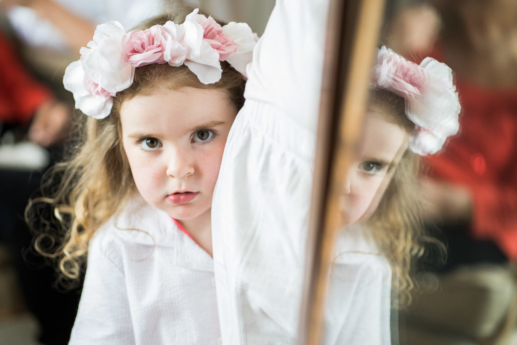 Shy flower girl wearing pink floral headband peering around large floor mirror