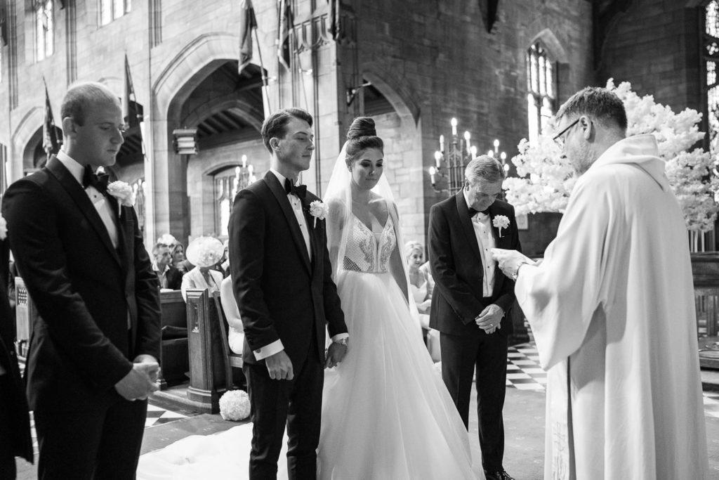 Bride and groom stood hand in hand and heads bowed before the vicar as prayers are spoken