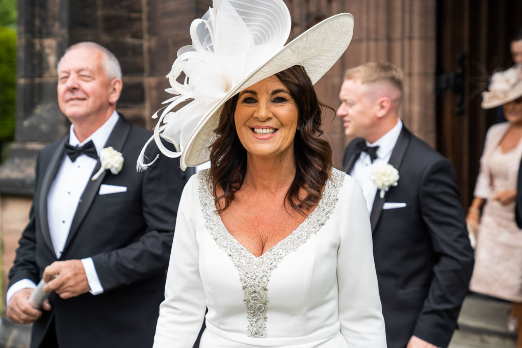 Mother of the bride smiling as she leaves the church wearing white outfit and large white hat