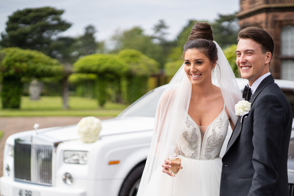 Bride and groom posed in front of their wedding car holding Prosecco while bouquet sits on car bonnet