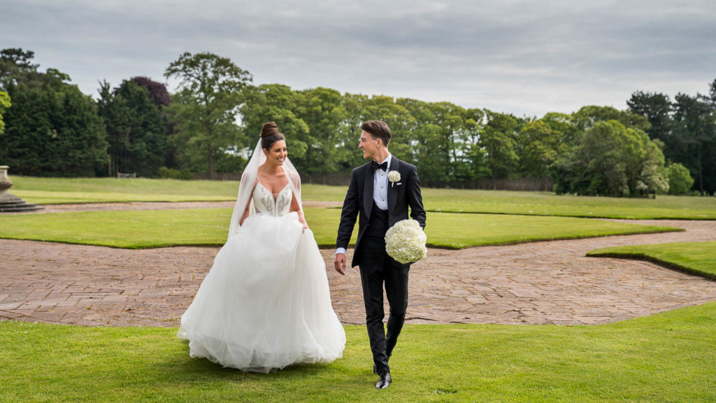 Groom walking with the bouquet as his brides lifts her dress to walk across the lawns