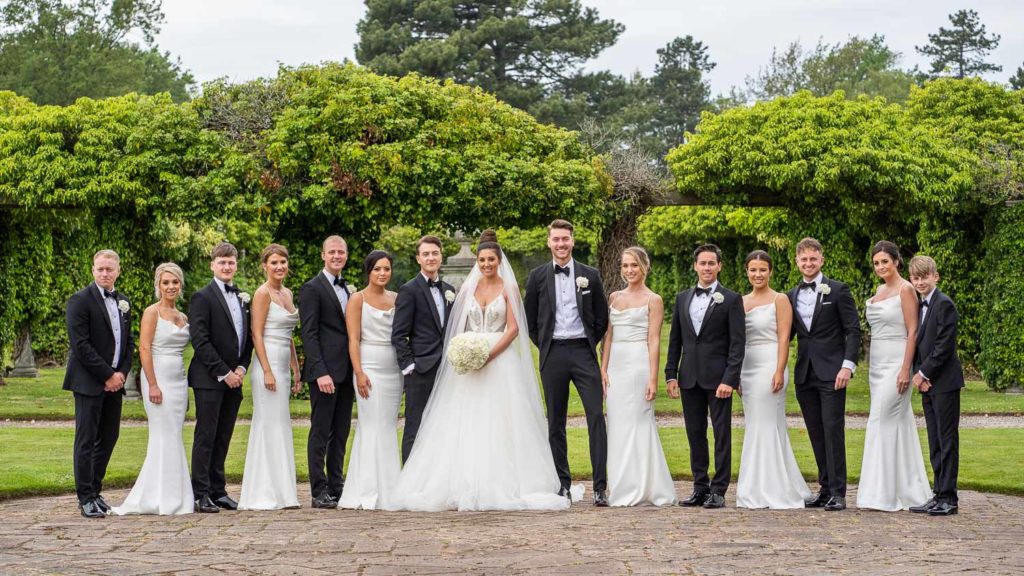 Full bridal party comprising bride and groom with seven groomsmen and six bridesmaids all wearing black and white