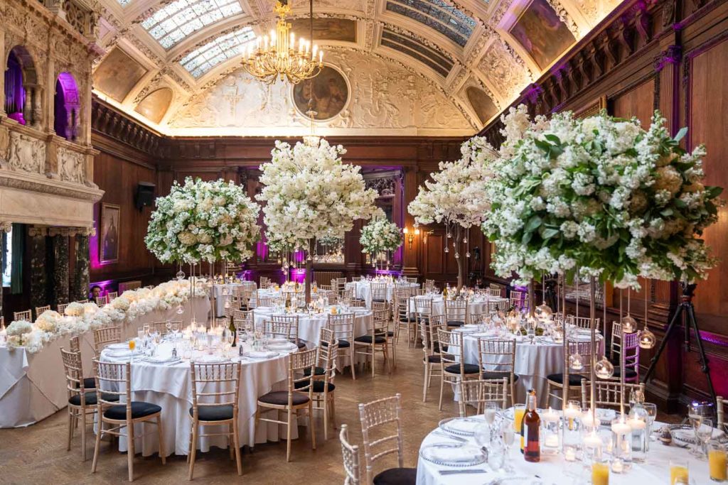 Wedding breakfast room with large hero centrepieces of white blooms and purple uplighters on the wooden walls