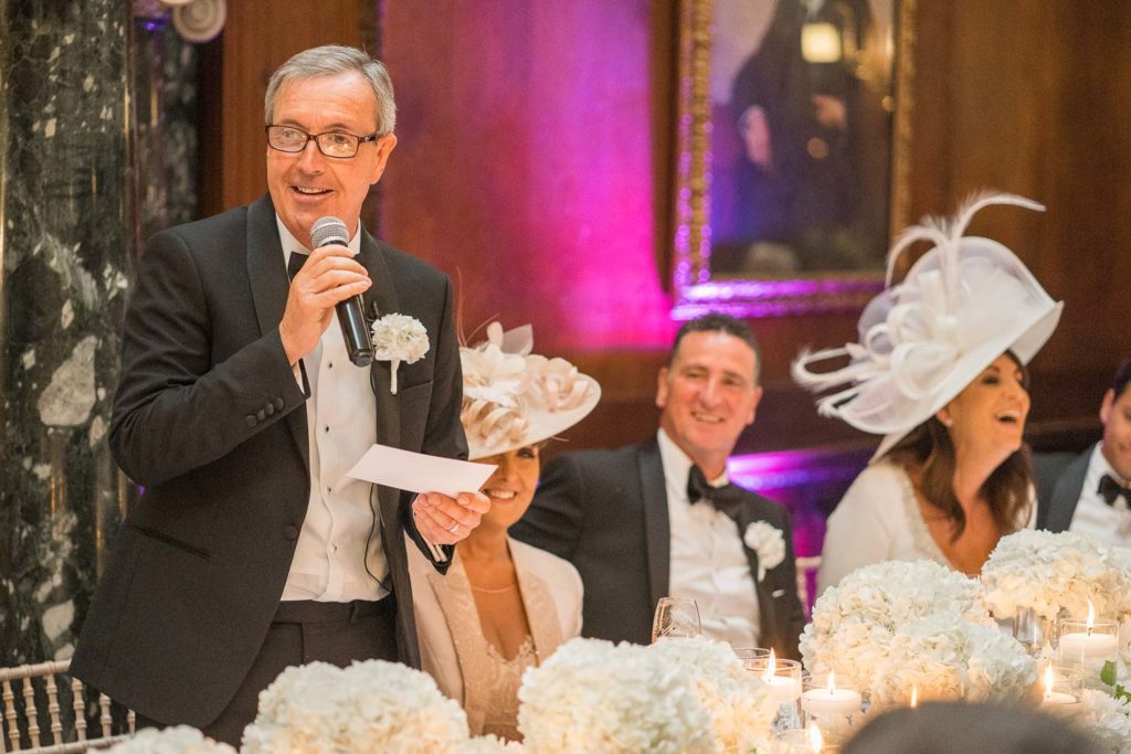 Father of the bride beginning the wedding speeches with guests on the top table laughing