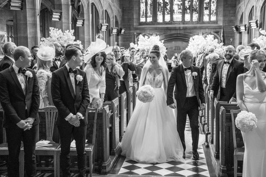 Bride and her father walking down the aisle, groom looks on, emotional, with all wedding guests smiling and bridesmaid wiping a tear